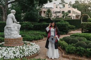 Graduate in cap and gown poses in formal garden with white statue and manicured hedges during outdoor graduation photo session.