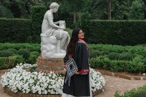 Graduate in cap and gown poses beside a white marble statue surrounded by white flowers in a formal garden.
