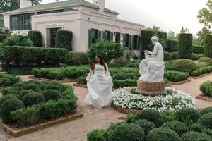 Bride in white gown walks through formal garden with sculpted hedges and stone statue near elegant estate.