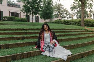 A smiling graduate in cap and gown sits on green terraced steps outdoors, holding a crochet piece.