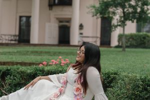 Woman in floral white dress sitting gracefully on green lawn beside blooming roses near elegant building.