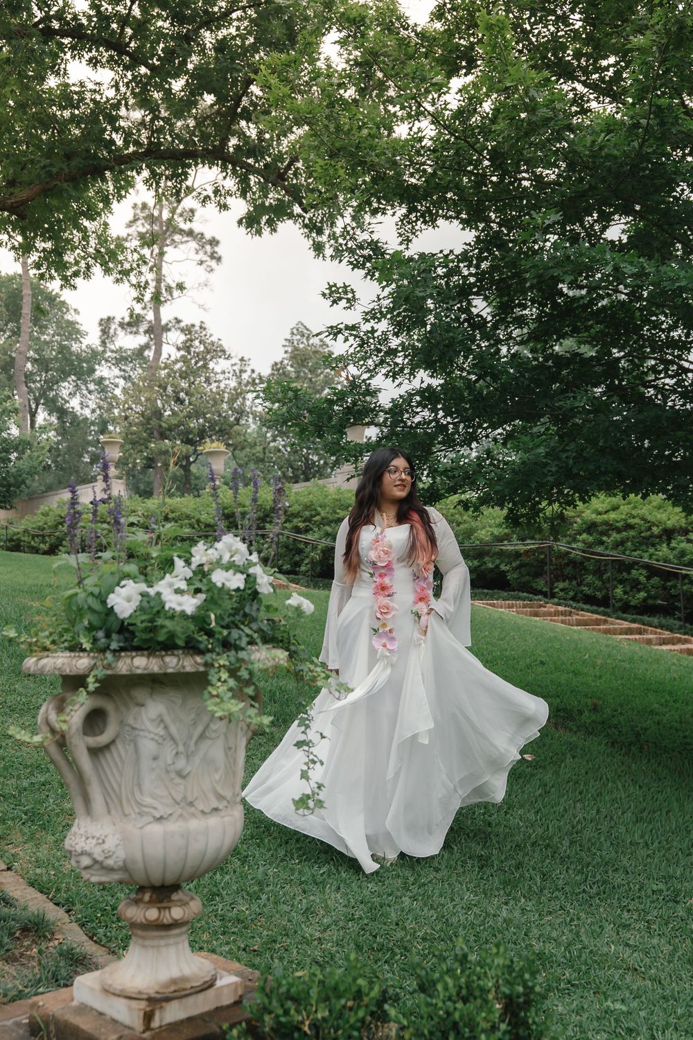 Bride in white floral gown twirling in lush garden beside stone urn with white flowers and green trees.