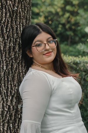 Young woman with glasses and gold necklace in white top leaning against tree in lush green garden.
