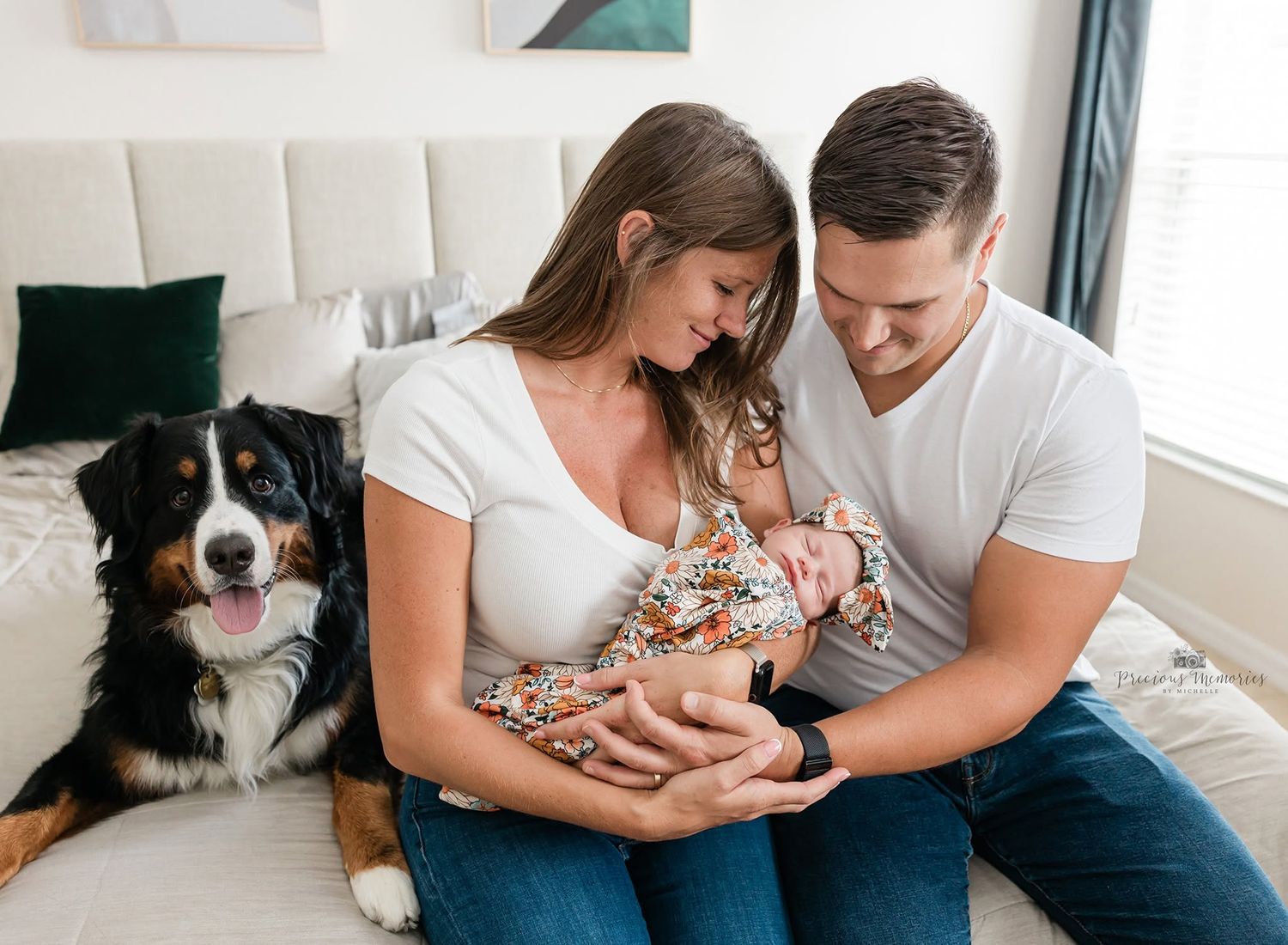 A couple sits on a bed holding their newborn baby, with a Bernese Mountain Dog beside them in a bright room.
