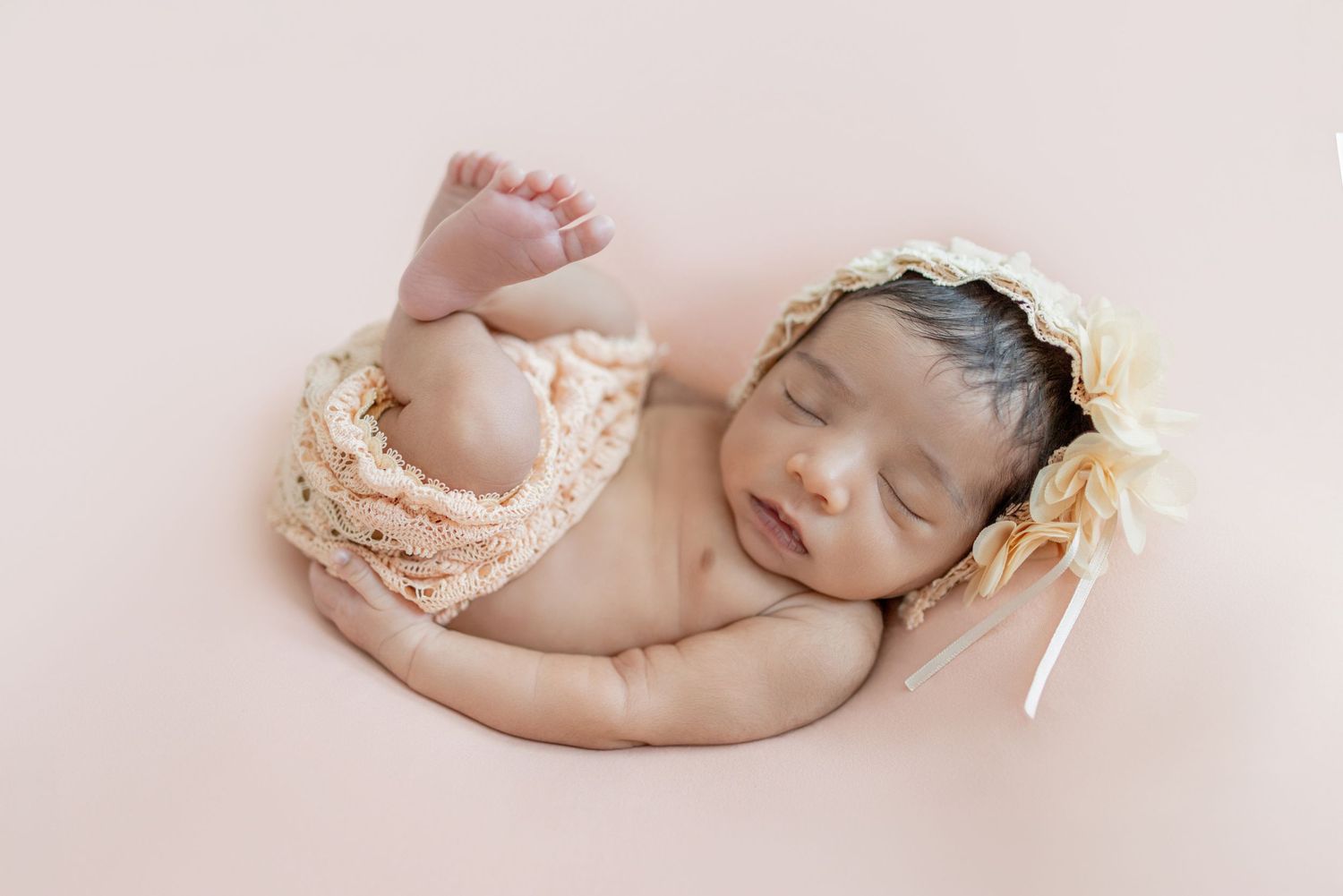 Sleeping newborn baby in peach knit wrap and floral bonnet posed on soft pink background.