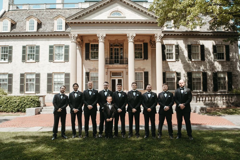 Groom and groomsmen in black tuxedos with bow ties posing in front of an elegant colonial mansion.