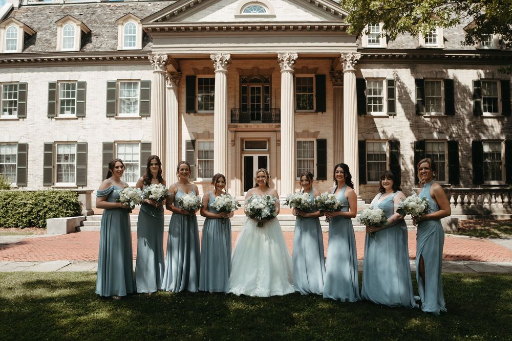 Bride and bridesmaids in sage green gowns holding white bouquets pose in front of an elegant colonial mansion.