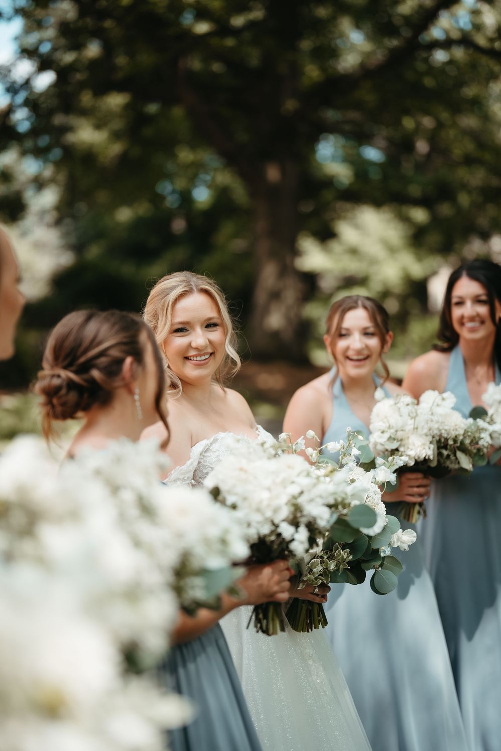 Bride in white gown smiles holding white floral bouquet surrounded by bridesmaids in light blue dresses outdoors.