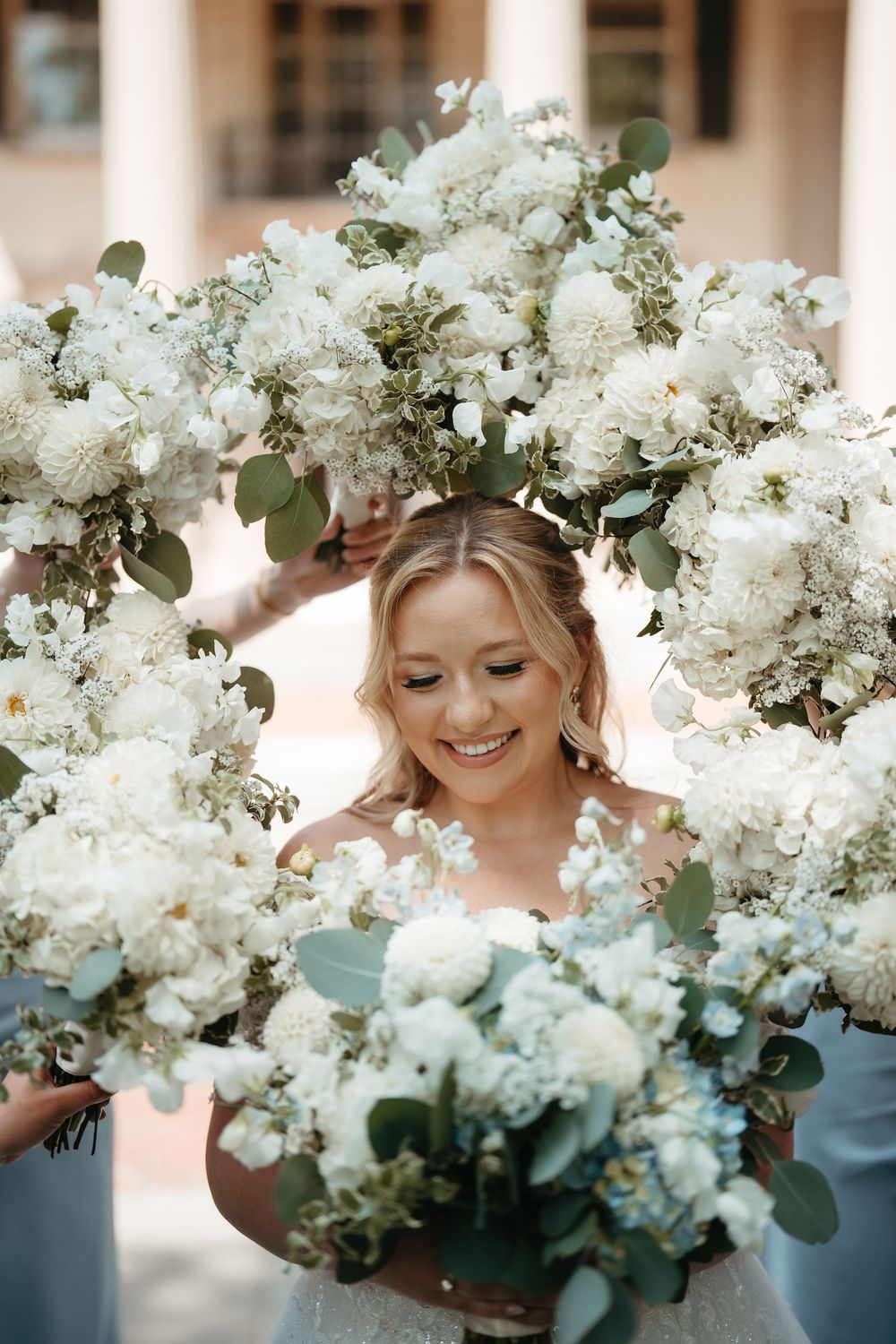 Smiling bride framed by white floral bouquets held by bridesmaids in blue dresses at outdoor wedding.