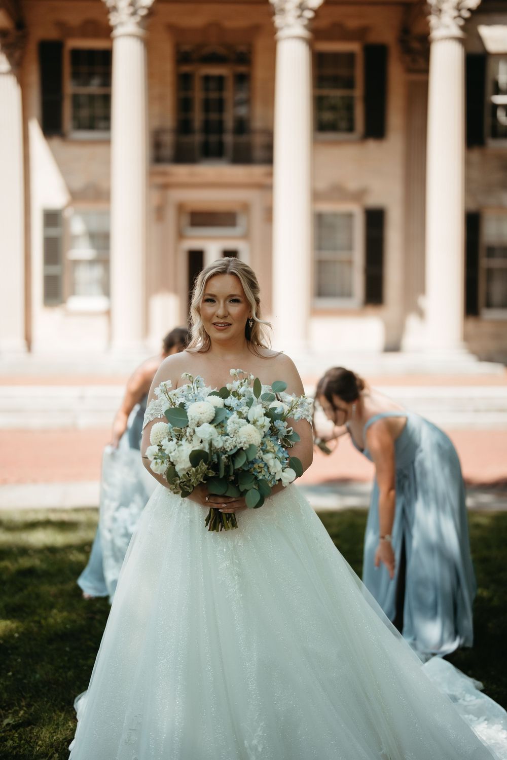 Bride in white ball gown holds blue and white bouquet before a grand columned mansion with bridesmaids in blue.