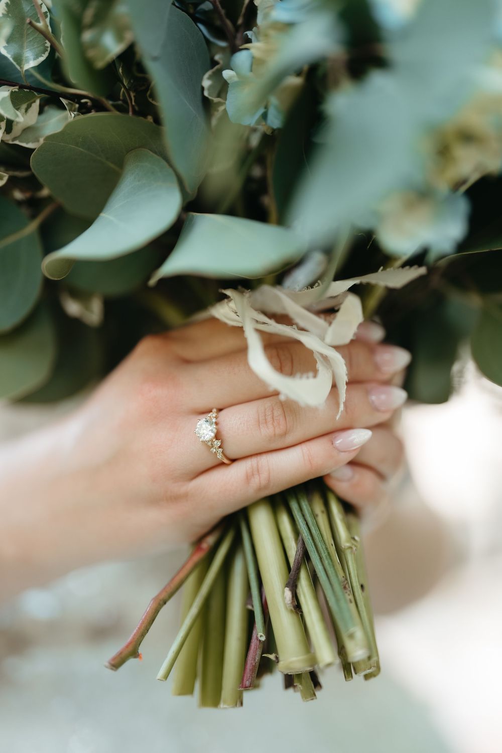 Close-up of bride's hand holding eucalyptus bouquet with diamond engagement ring and nude manicure.
