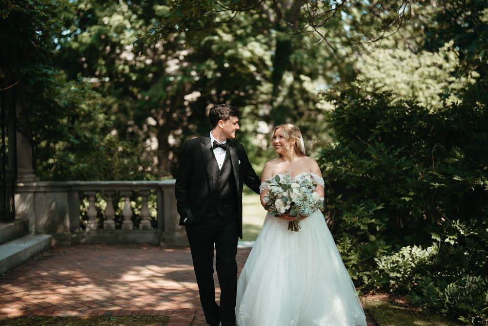 Bride and groom walking hand in hand in lush garden, she in off-shoulder gown holding bouquet, he in classic black tuxedo.