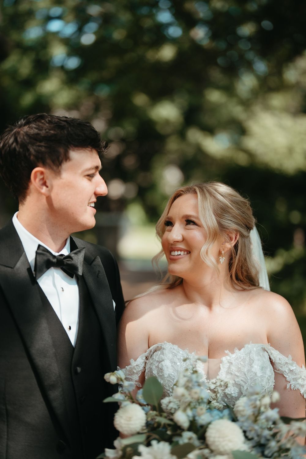 A smiling bride in a floral gown holds a bouquet while gazing at her groom in a black tuxedo outdoors.