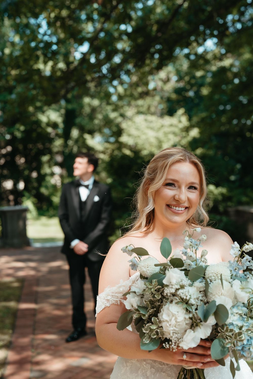 Smiling bride holding white and eucalyptus bouquet outdoors, groom in tuxedo standing in background on brick path.