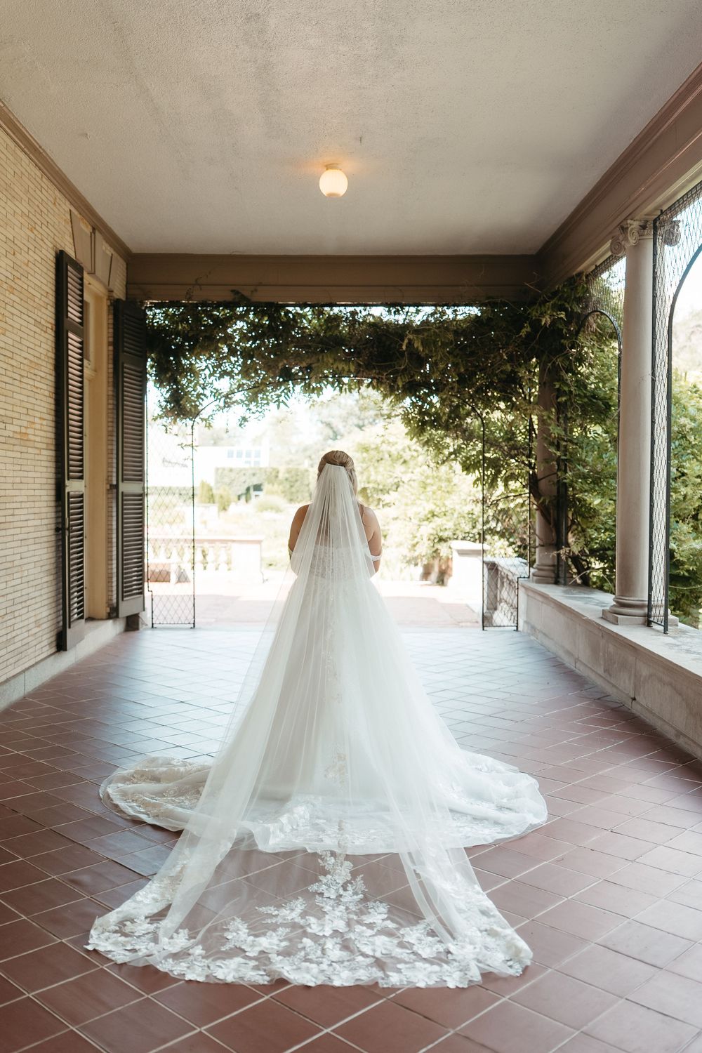 Bride in white gown with long lace-trimmed cathedral veil stands on elegant tiled portico overlooking garden.