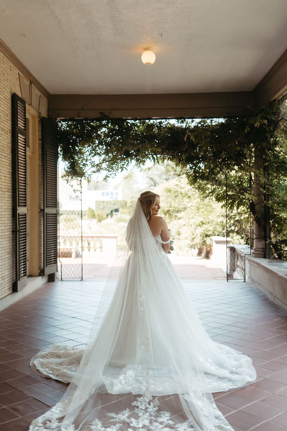 Bride in strapless lace wedding gown with long cathedral veil stands on elegant venue porch surrounded by lush greenery.