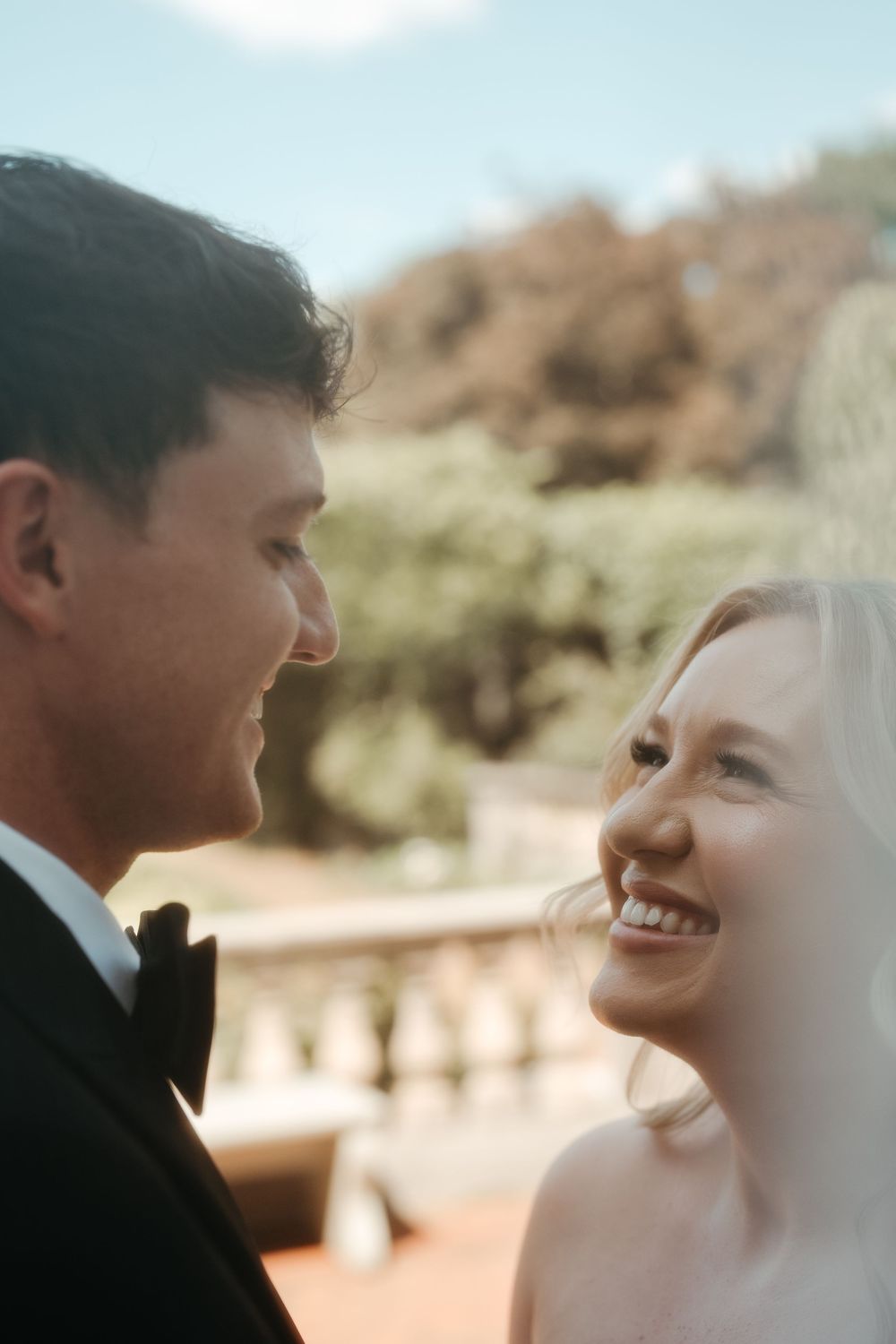 A bride and groom share a joyful moment, smiling at each other outdoors on their wedding day.