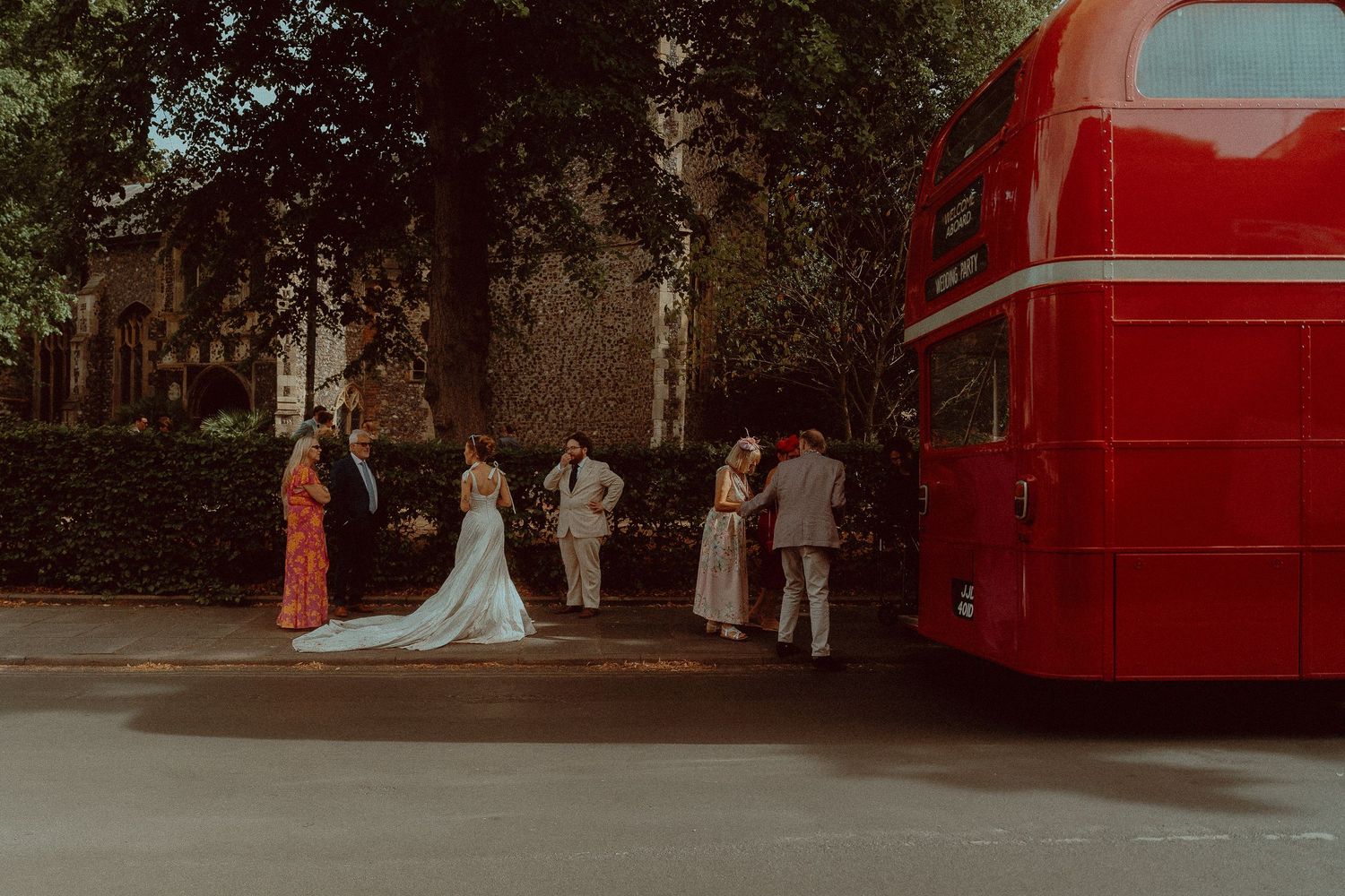 Wedding party boarding a red double-decker London bus, bride in white gown, guests in formal attire, tree-lined street.