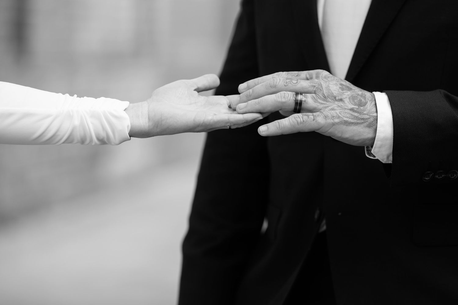 Black and white photo of a couple exchanging wedding rings, groom wearing a suit with tattooed hands.
