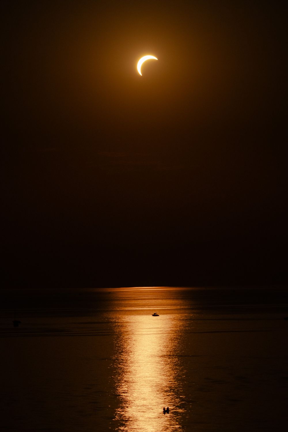 A partial solar eclipse glows golden over a calm ocean at dusk, with a boat silhouetted on the reflective water below.