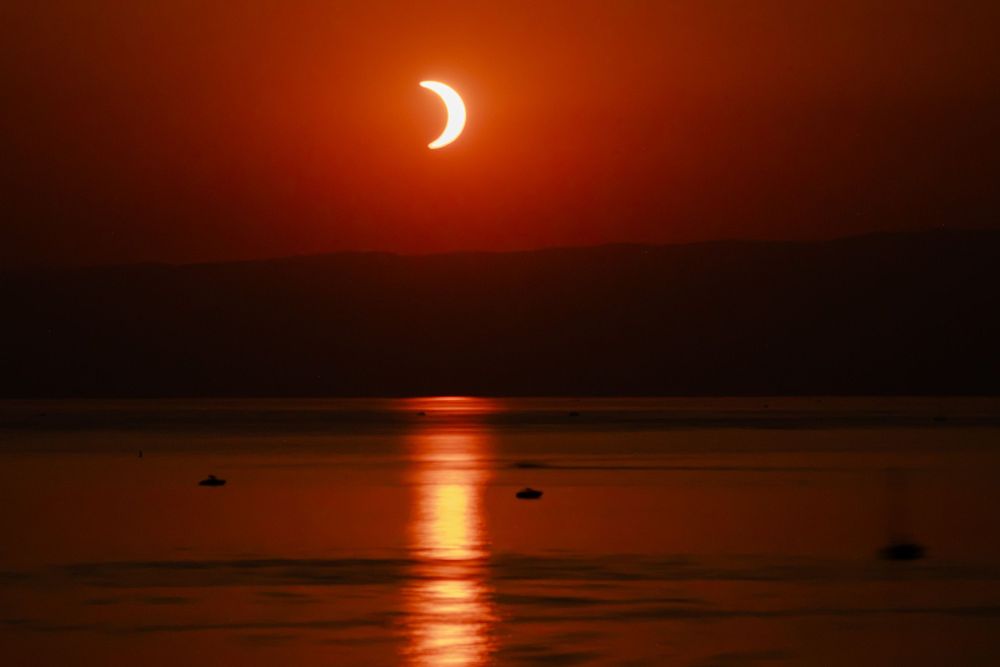 Partial solar eclipse over a calm ocean at sunset with vivid red and orange sky reflecting on the water.