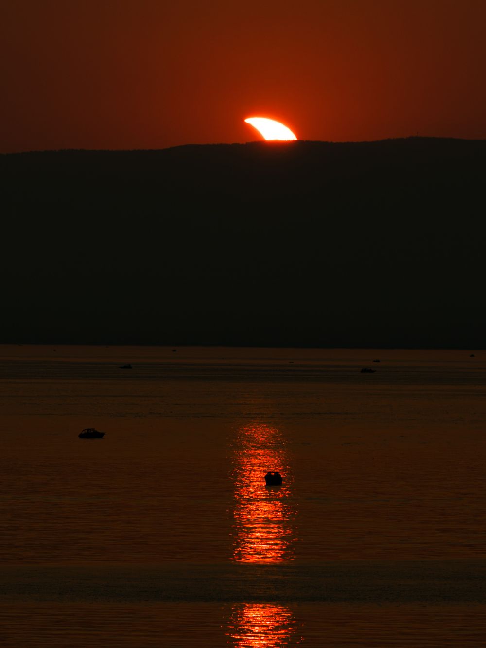A stunning red sunset over a calm lake with the sun setting behind a dark hillside, reflecting orange light on the water.
