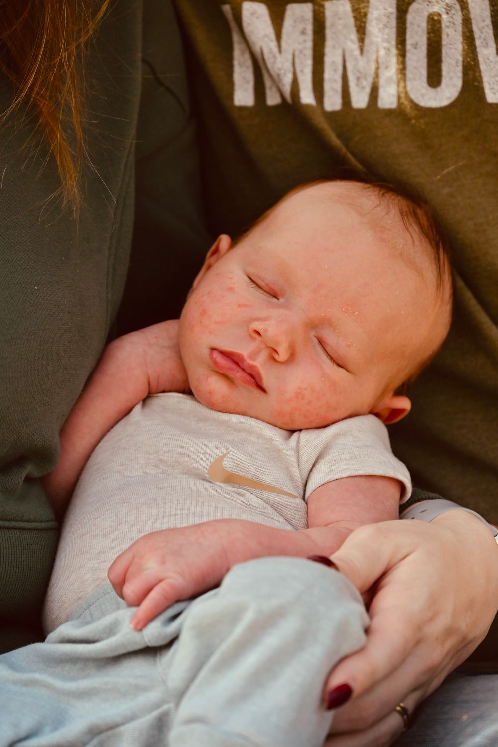 Newborn baby sleeping peacefully in mother's arms, wearing a white Nike onesie with rosy skin.
