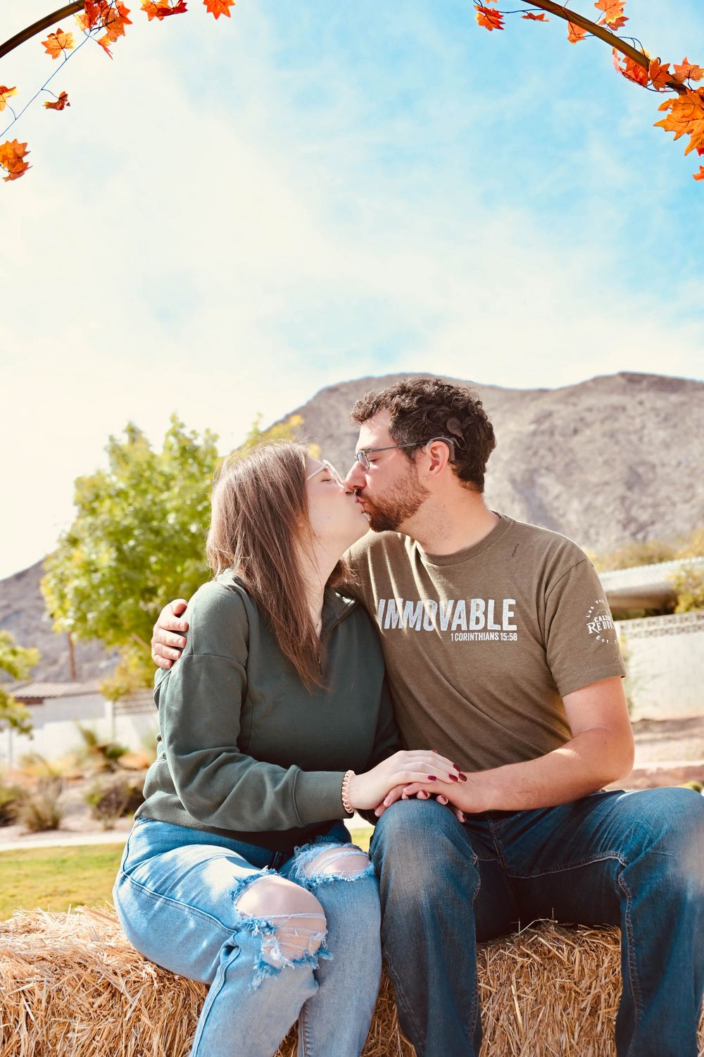 Couple sharing a romantic kiss on a hay bale with autumn leaves and mountain backdrop.