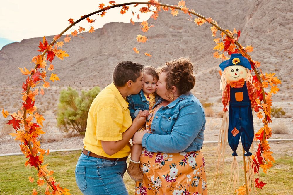 Family sharing a kiss under a fall leaf arch with a scarecrow decoration and desert mountains in the background.