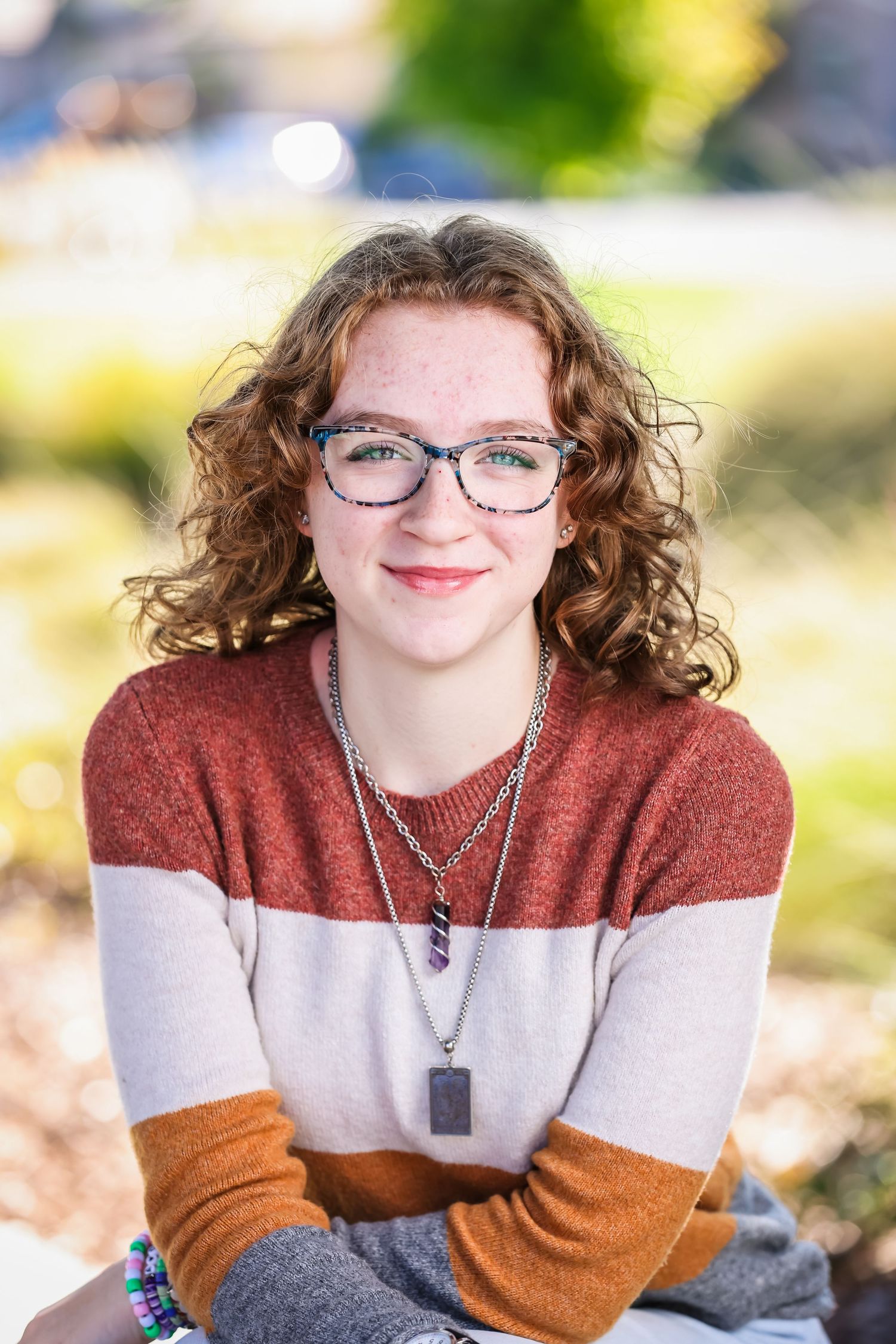Young woman with curly red hair and blue glasses wearing a striped sweater, smiling outdoors in natural light.