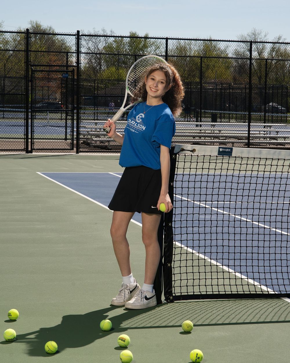 Young female tennis player posing on court holding racket near net surrounded by tennis balls wearing blue shirt and black skirt.