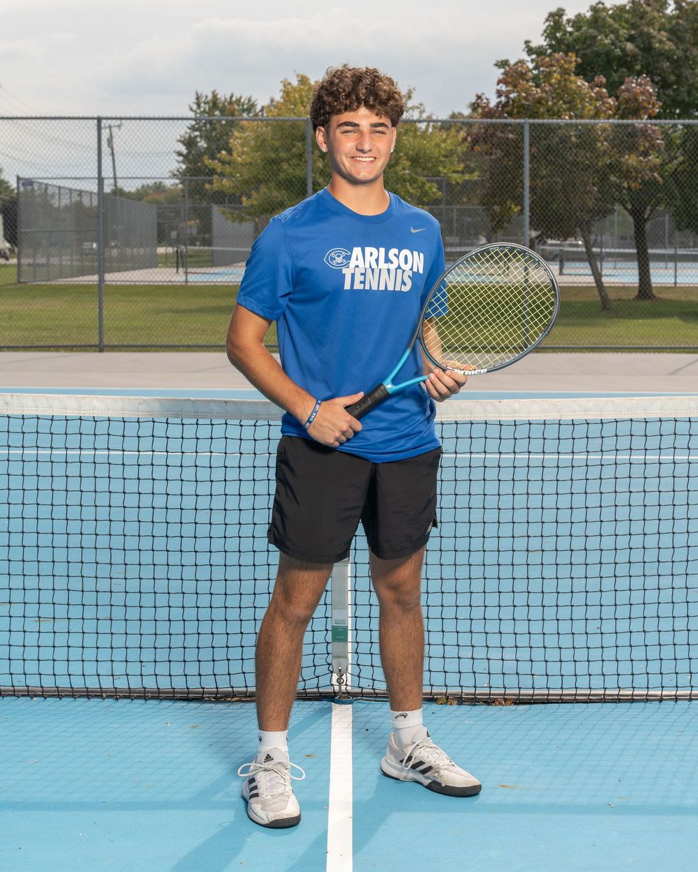 Young male tennis player in blue Arlson Tennis shirt holds racket while posing at net on outdoor court.