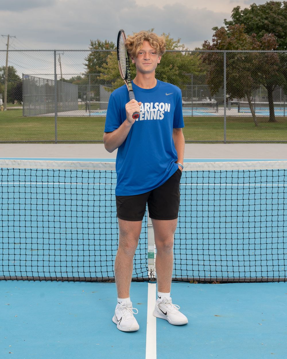 Young tennis player in blue Carlson Tennis shirt holds racket at net on outdoor blue court.