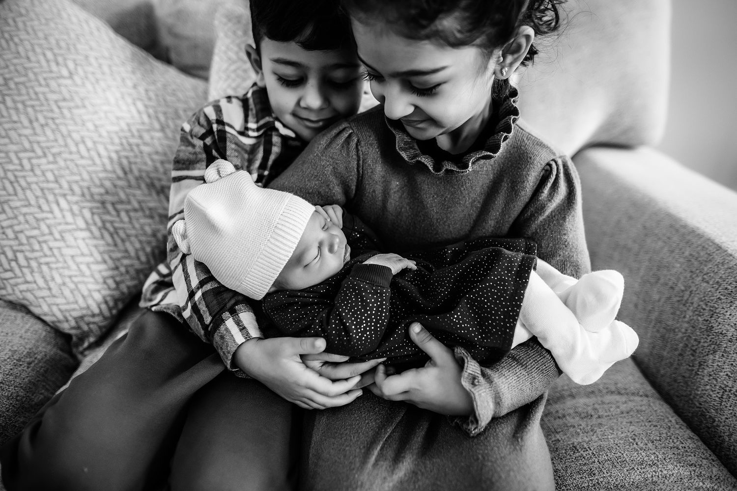 Black and white photo of two young siblings lovingly holding a sleeping newborn baby on a couch.