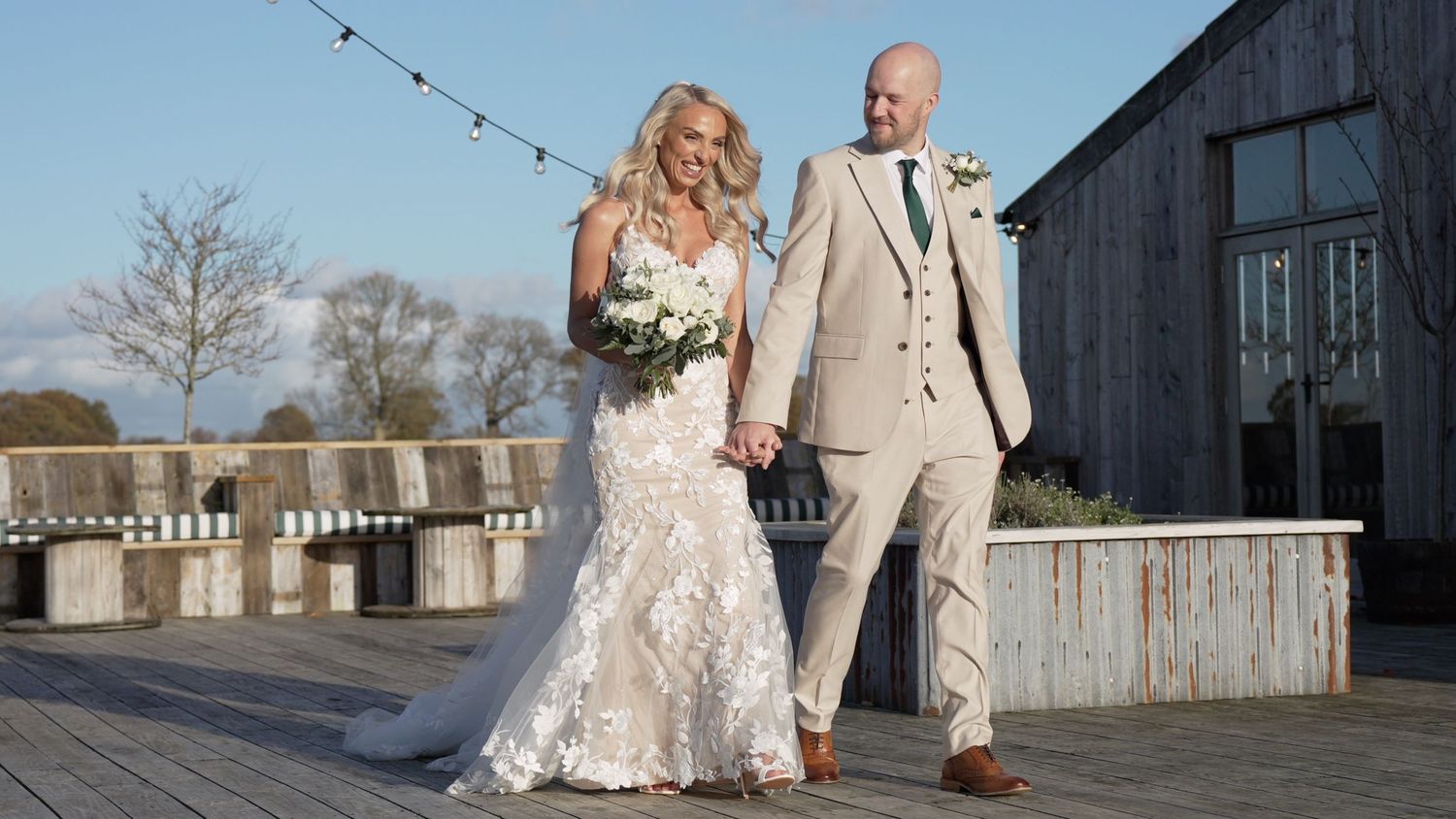 A bride and groom hold hands on a wooden deck, she in a lace gown, he in a beige suit, on their wedding day.