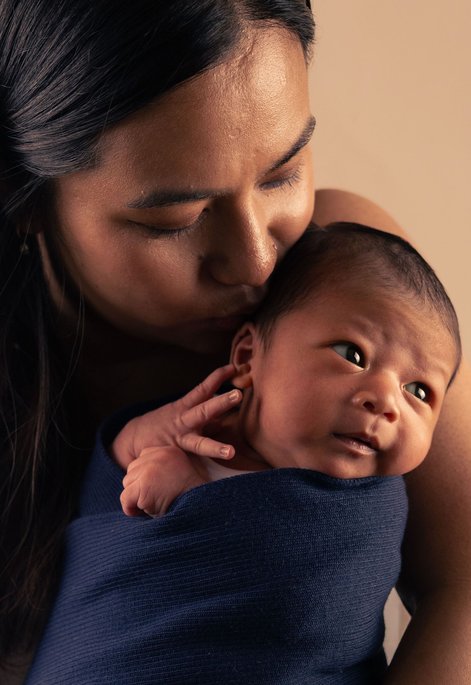 A mother tenderly kisses her newborn baby wrapped in a navy swaddle blanket against a warm beige background.