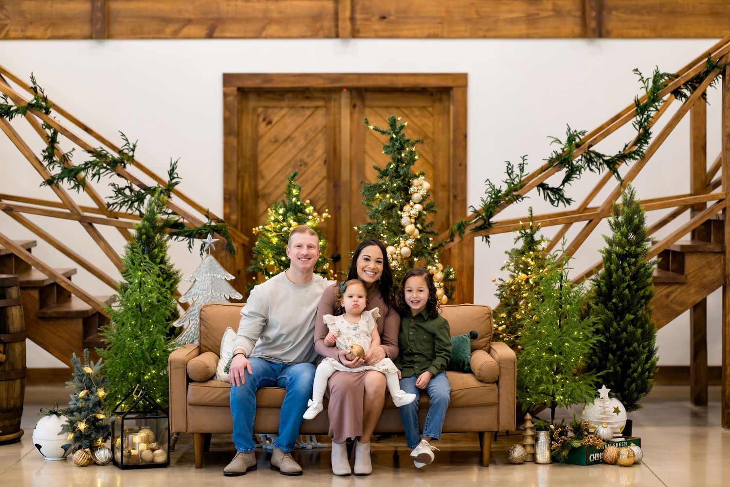 Family of four poses on a couch surrounded by Christmas trees and holiday decor in a festive indoor setting.