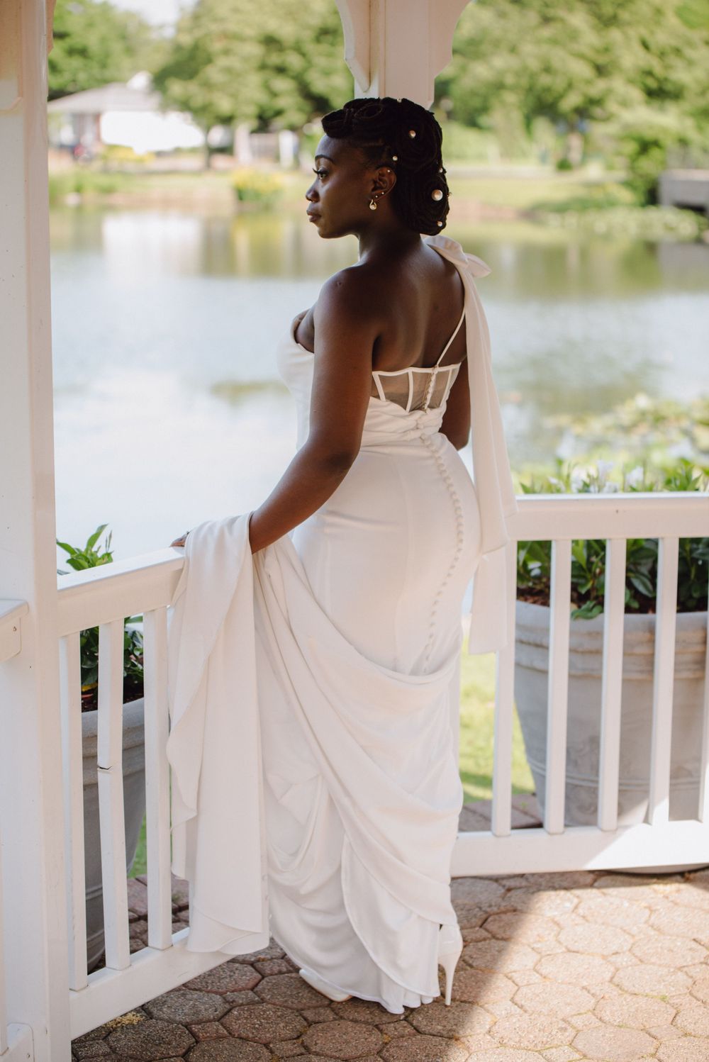 A bride in a white dress stands on a gazebo overlooking a peaceful lake.