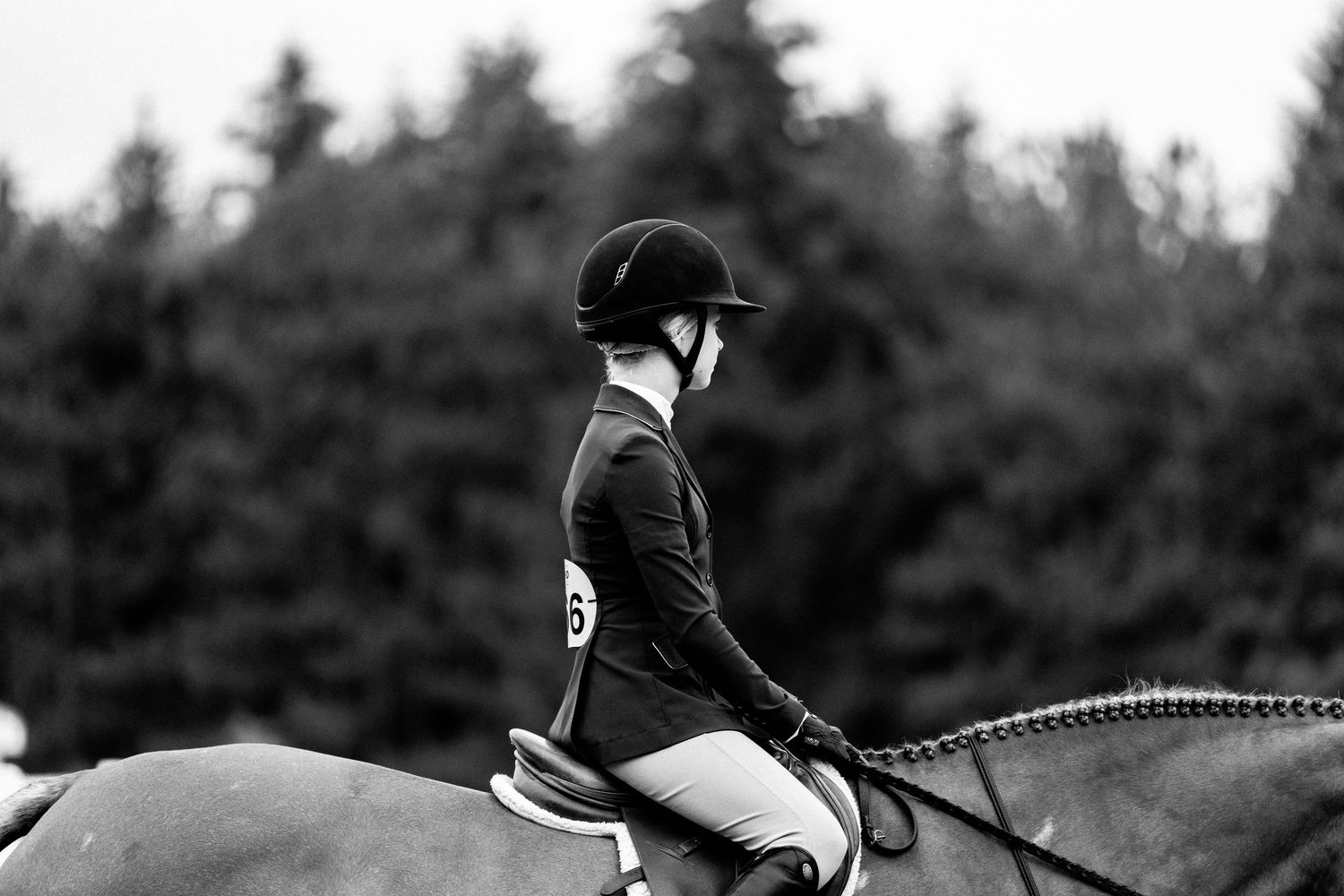 Black and white photo of an equestrian rider in helmet and jacket riding a horse with trees in background.