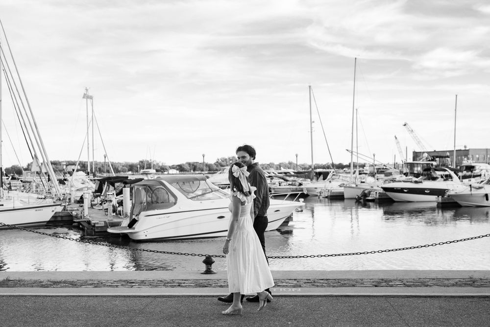 Black and white photo of a couple embracing at a marina with sailboats in the background on a cloudy day.