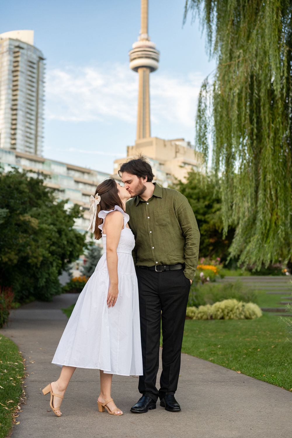 Couple sharing a kiss in a Toronto park with the CN Tower visible in the background on a sunny day.