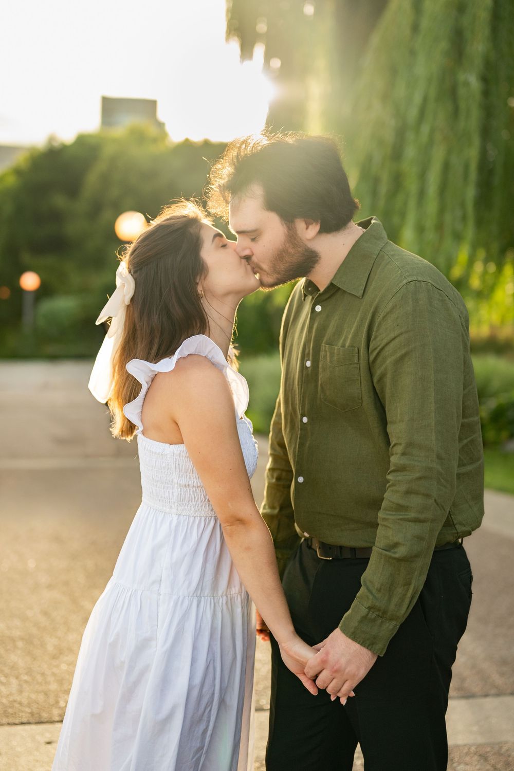 Couple kissing at golden hour in a park, woman in white dress holding hands with man in green shirt.
