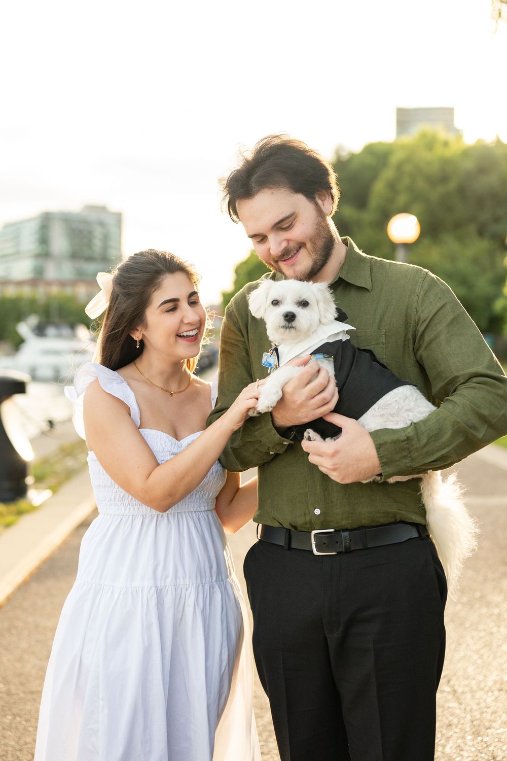 A smiling couple in elegant attire holds a small white dog dressed in a tuxedo outdoors at golden hour.