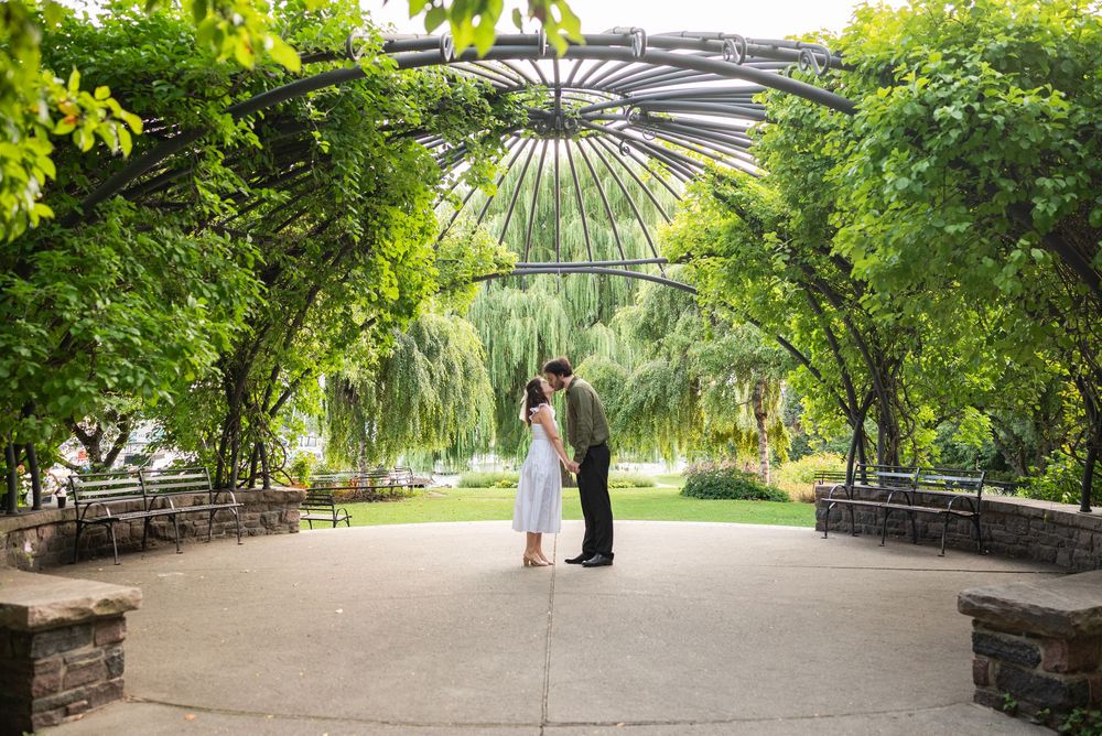 Couple sharing a romantic kiss under a lush green iron pergola arbor in a scenic outdoor garden park.