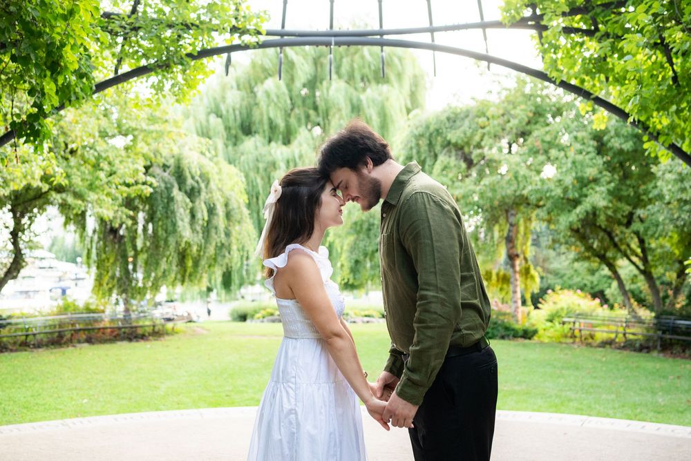 Romantic couple holding hands and touching foreheads under an arch in a lush green park setting.