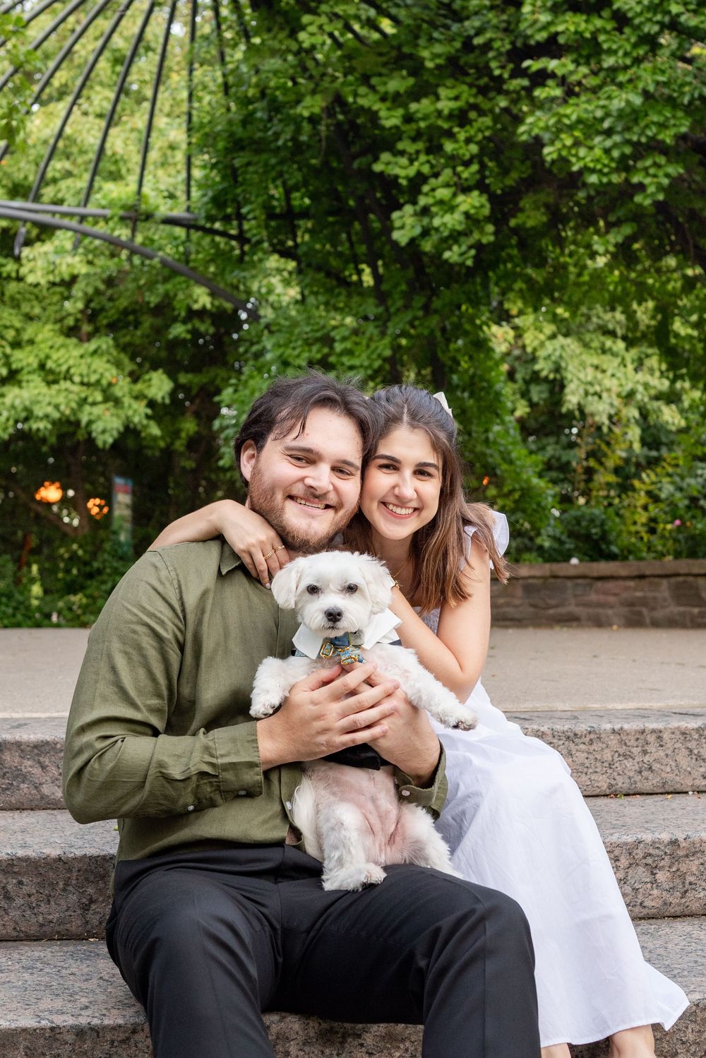 Smiling couple sitting on stone steps outdoors holding a small white dog, surrounded by lush green trees.