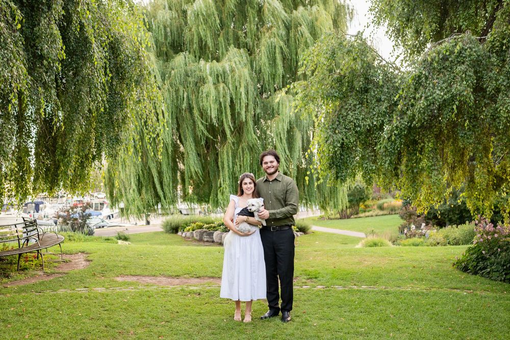 Happy couple standing together on green grass beneath a weeping willow tree in a scenic park setting.