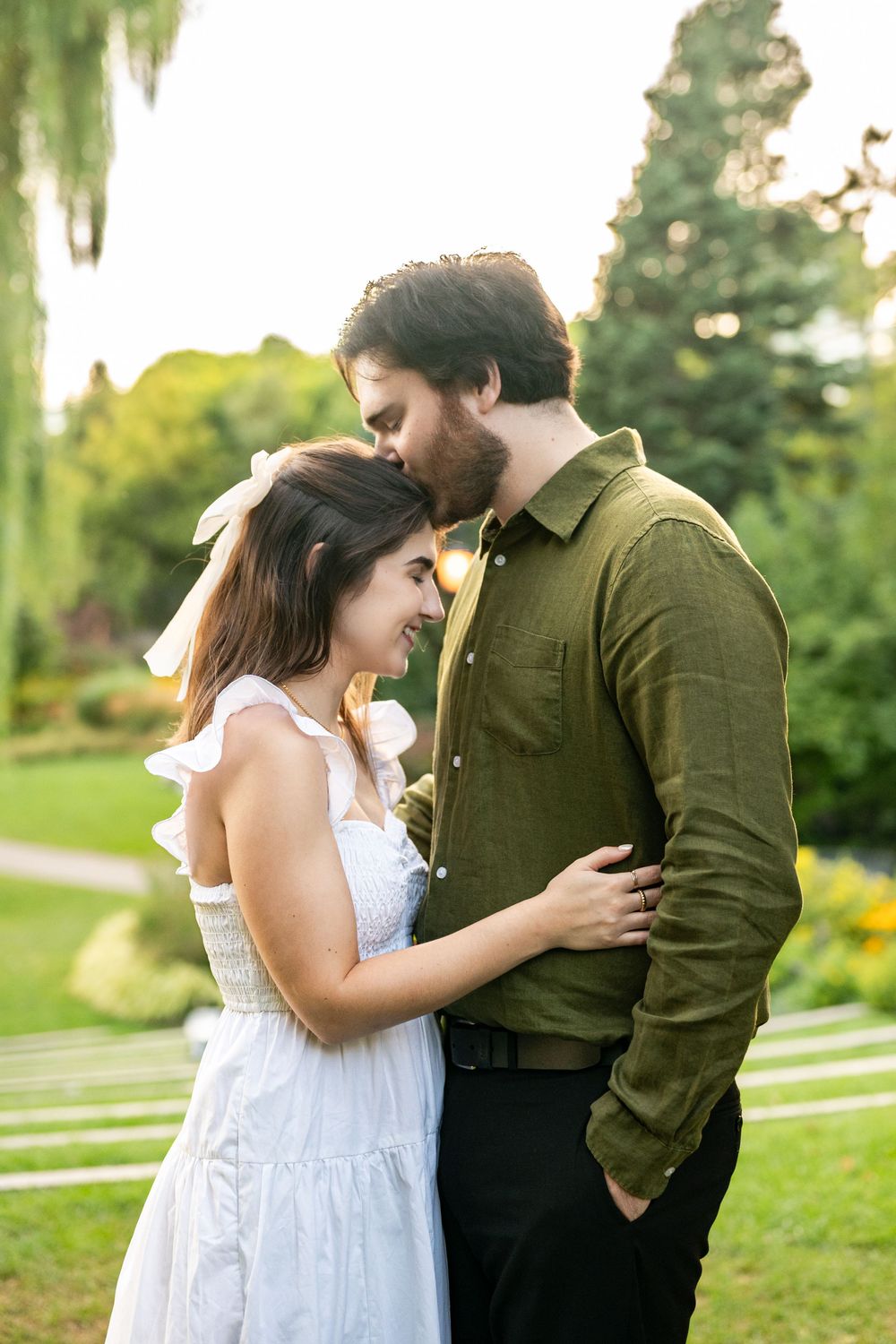 A couple embraces tenderly in a lush green garden, woman in white dress smiling up at bearded man in olive shirt.