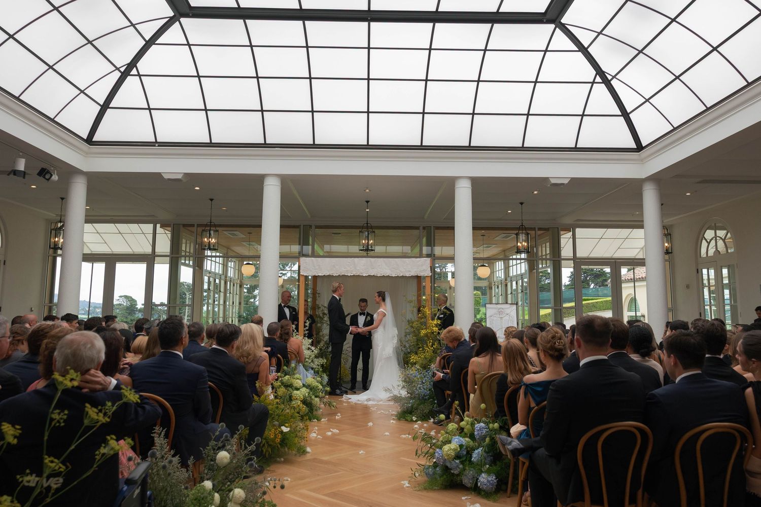 Indoor wedding ceremony in elegant glass-ceiling venue with floral arch, seated guests, and lush greenery lining the aisle.