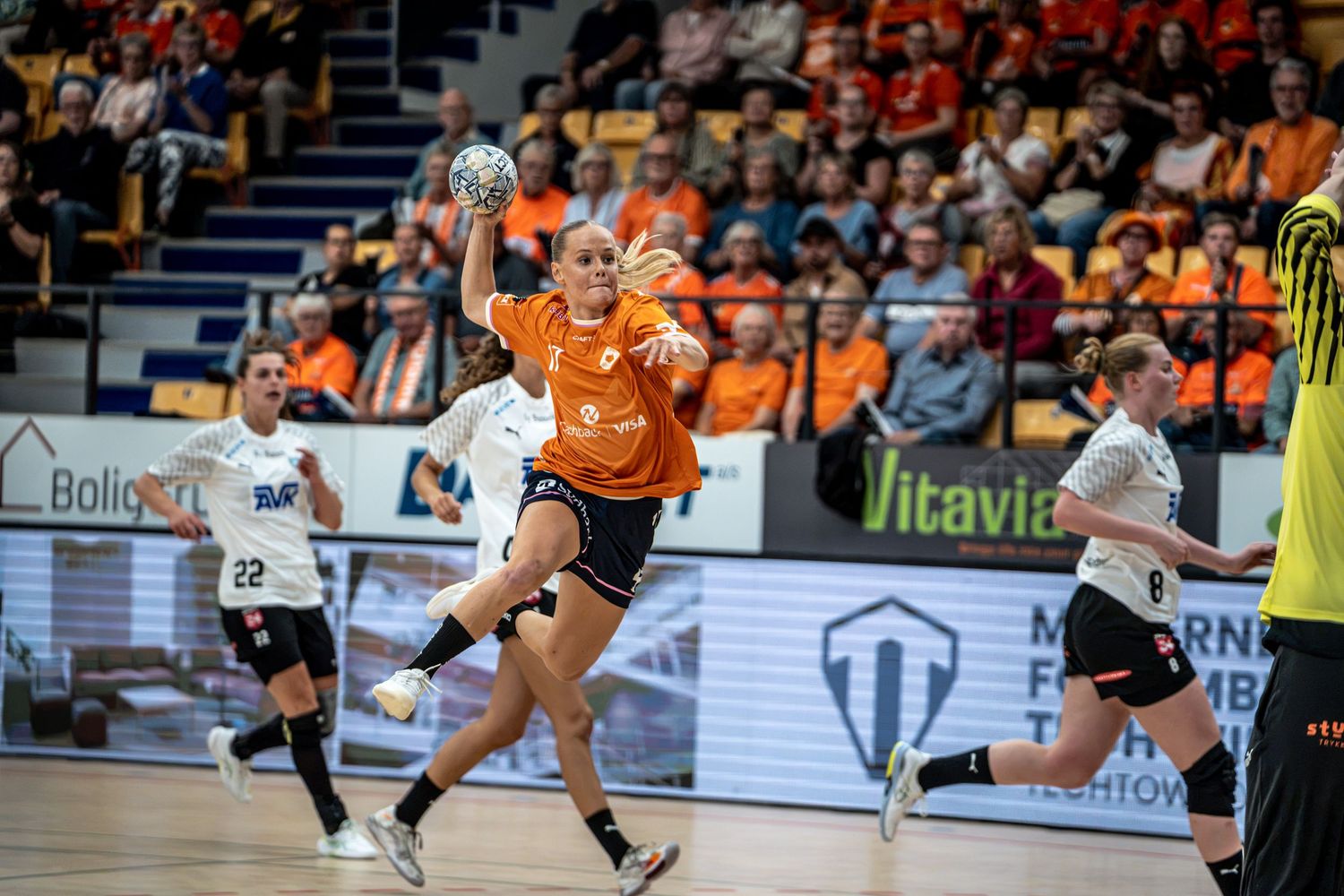 Female handball player in orange jersey leaping to throw ball during competitive indoor match.